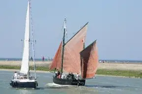 Voilier traditionnel à voiles ocre et voilier moderne à voile blanche naviguant côte à côte dans le chenal de l’Aa, devant la plage et les jetées de Gravelines.