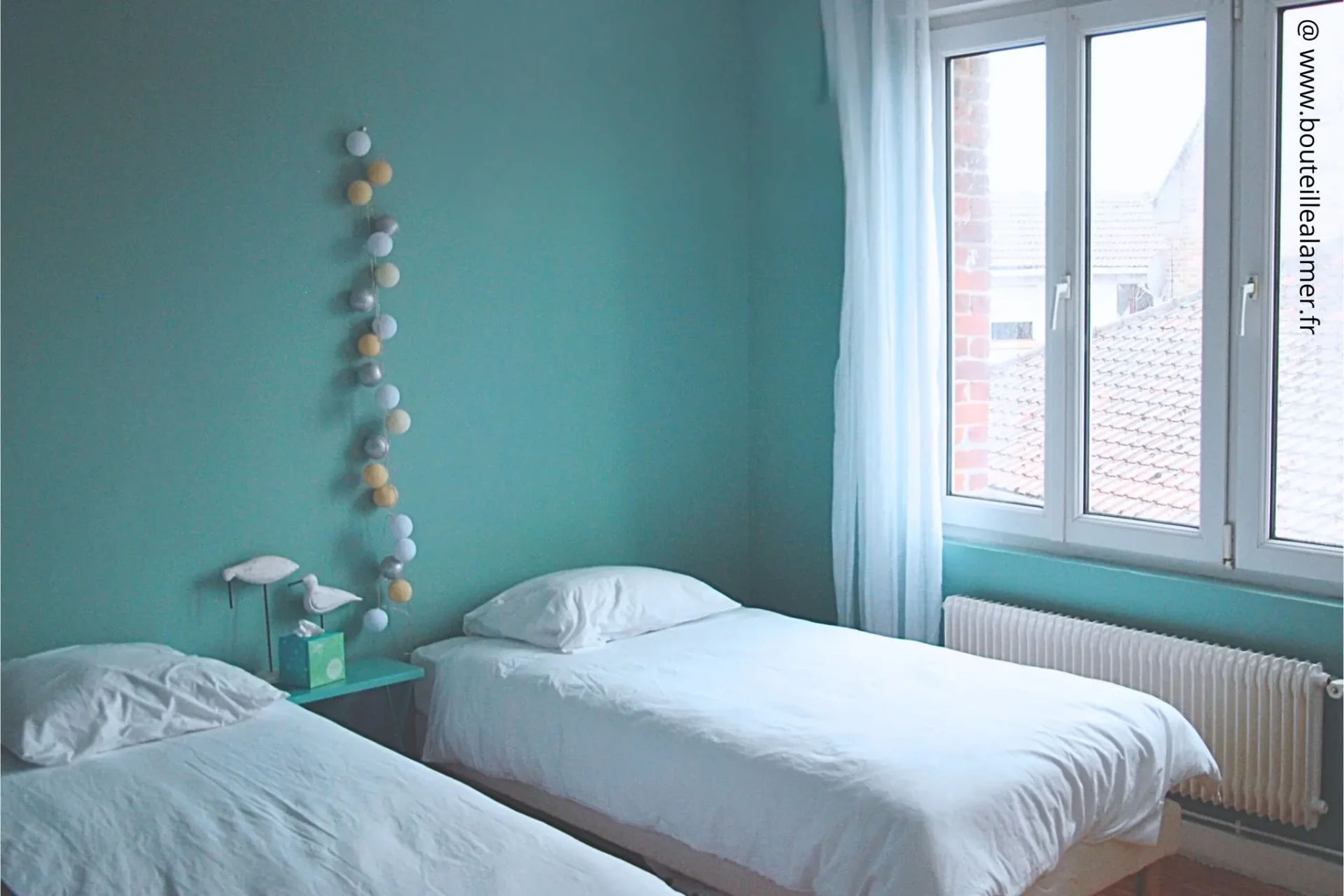 Chambre lumineuse avec deux lits simples, murs bleu-vert, grande fenêtre donnant sur le jardin côté bourg, gîte Bouteille à la Mer, Côte d’Opale, Gravelines, maison de vacances en location.