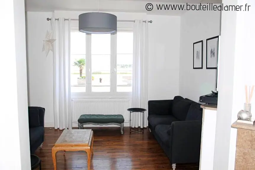 Salon lumineux avec deux canapés, grande fenêtre offrant une vue sur la mer et le passage des bateaux à marée haute, parquet en bois, gîte Bouteille à la Mer, Côte d’Opale, Gravelines.