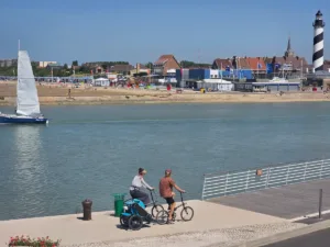 Vue sur le chenal de l’Aa à Gravelines avec un voilier, deux cyclistes sur la Vélomaritime (EuroVelo 4), les restaurants et maisons du front de mer, et le phare rayé noir et blanc de Gravelines sur la Côte d’Opale.